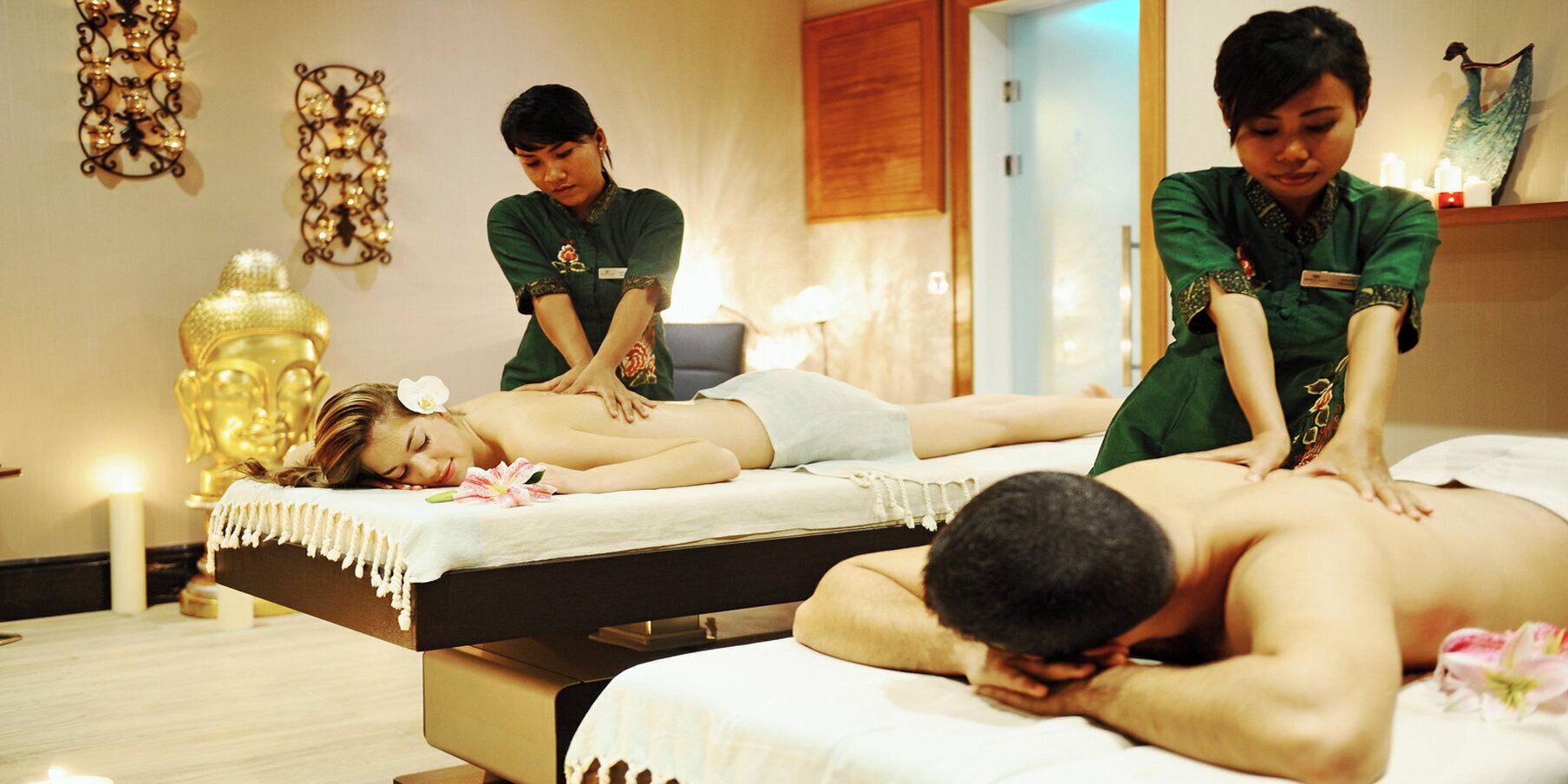 Un salón de terapia de masaje con dos clientes recibiendo tratamiento, un terapeuta atendiendo y una estatua de Buda al fondo.