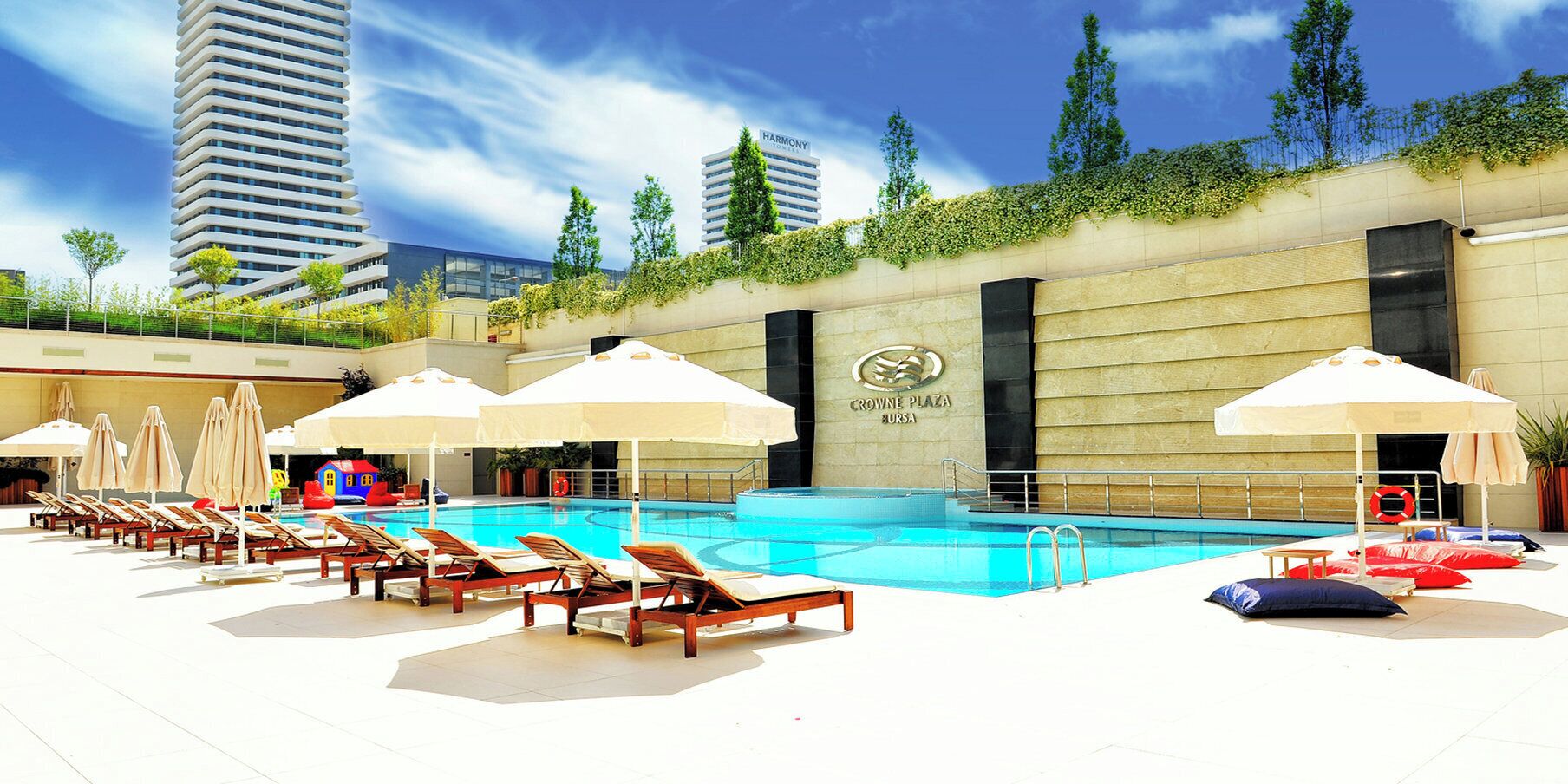 Zona de piscina con sillas de descanso y sombrillas, rodeada por un muro bajo con vegetación.