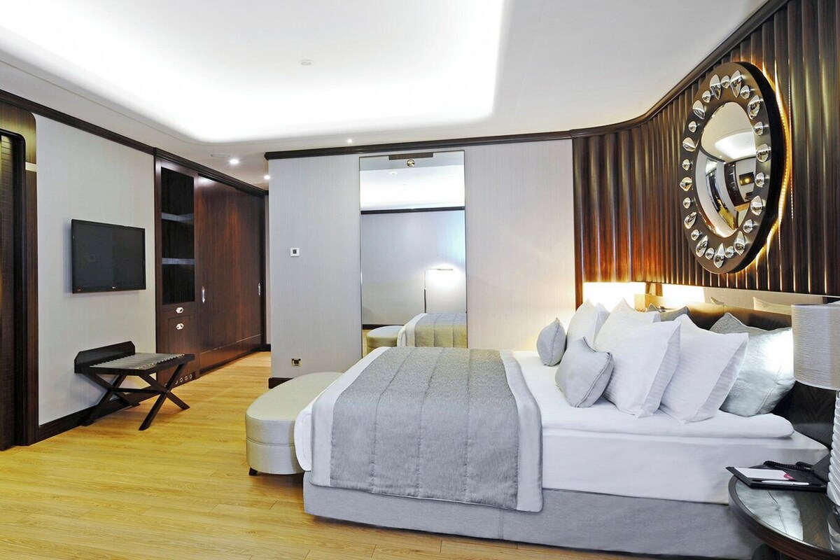 A modern bedroom with a large bed, a desk, a chair, a TV, and a mirror.