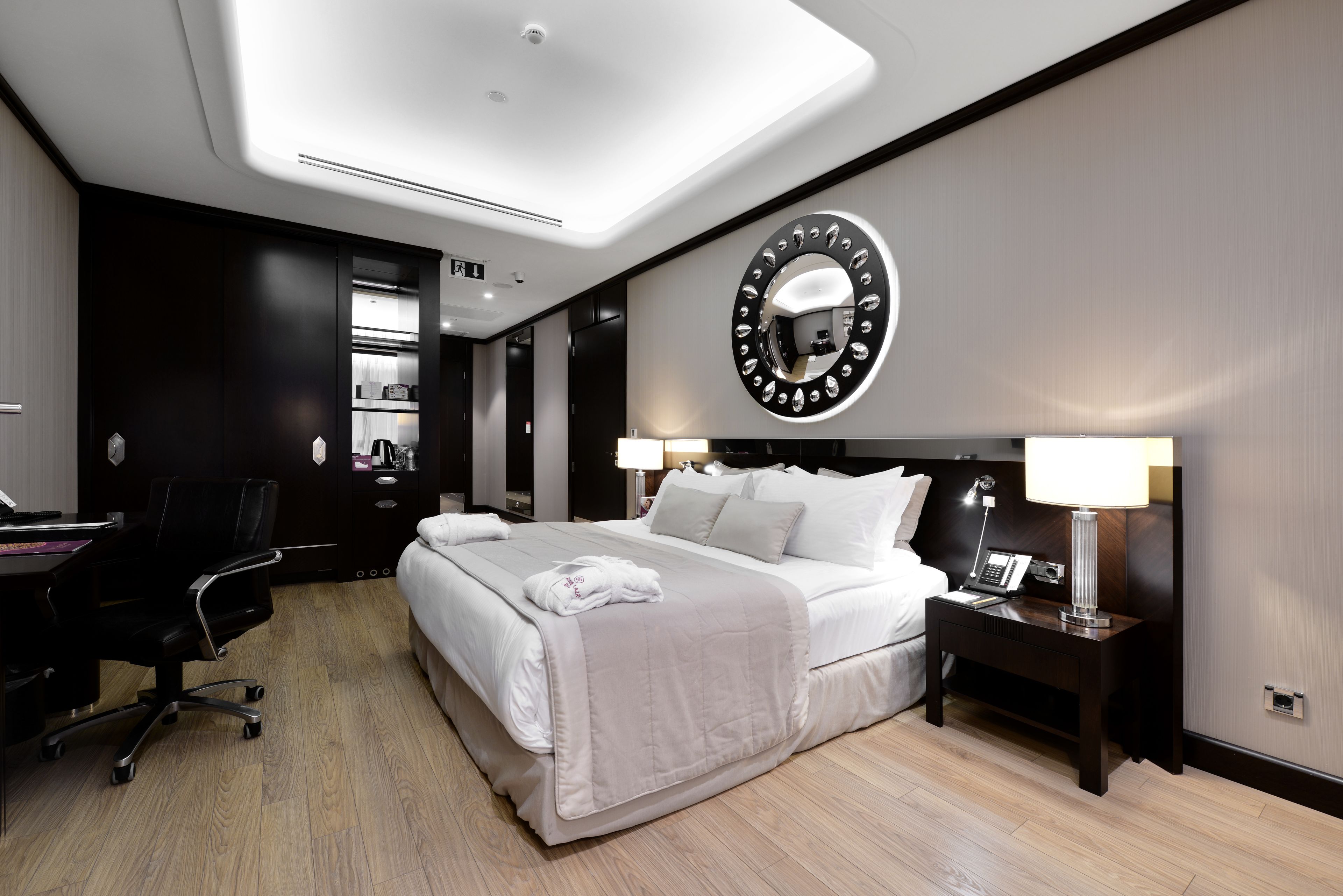 A modern hotel room with a large bed, a desk, and a chair. There is a round mirror on the wall and a nightstand with a lamp.