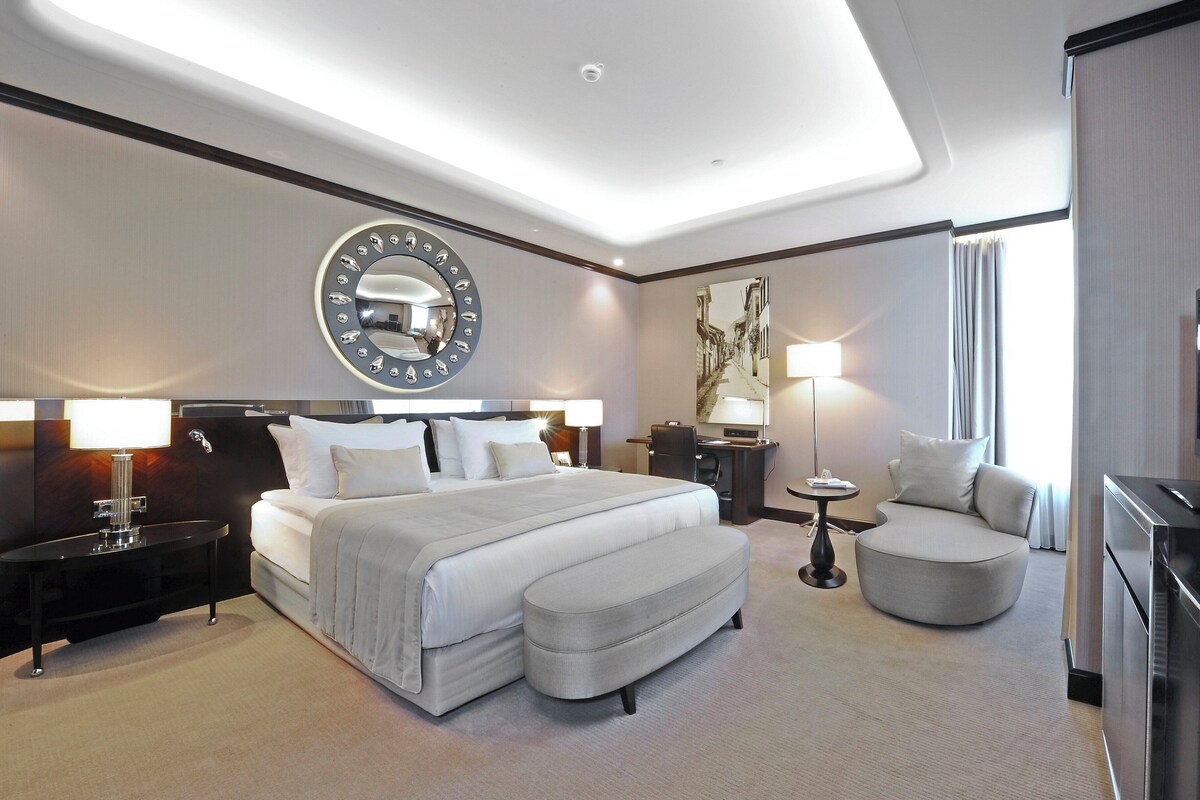 A hotel room with a large bed, a round mirror, a desk, a chair, a television, and a small table.
