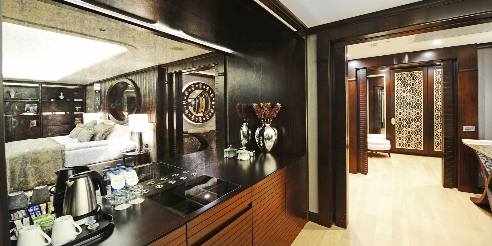 Habitación de hotel moderna con detalles en madera oscura, un espejo grande, un reloj redondo y una bandeja de desayuno con café y té.