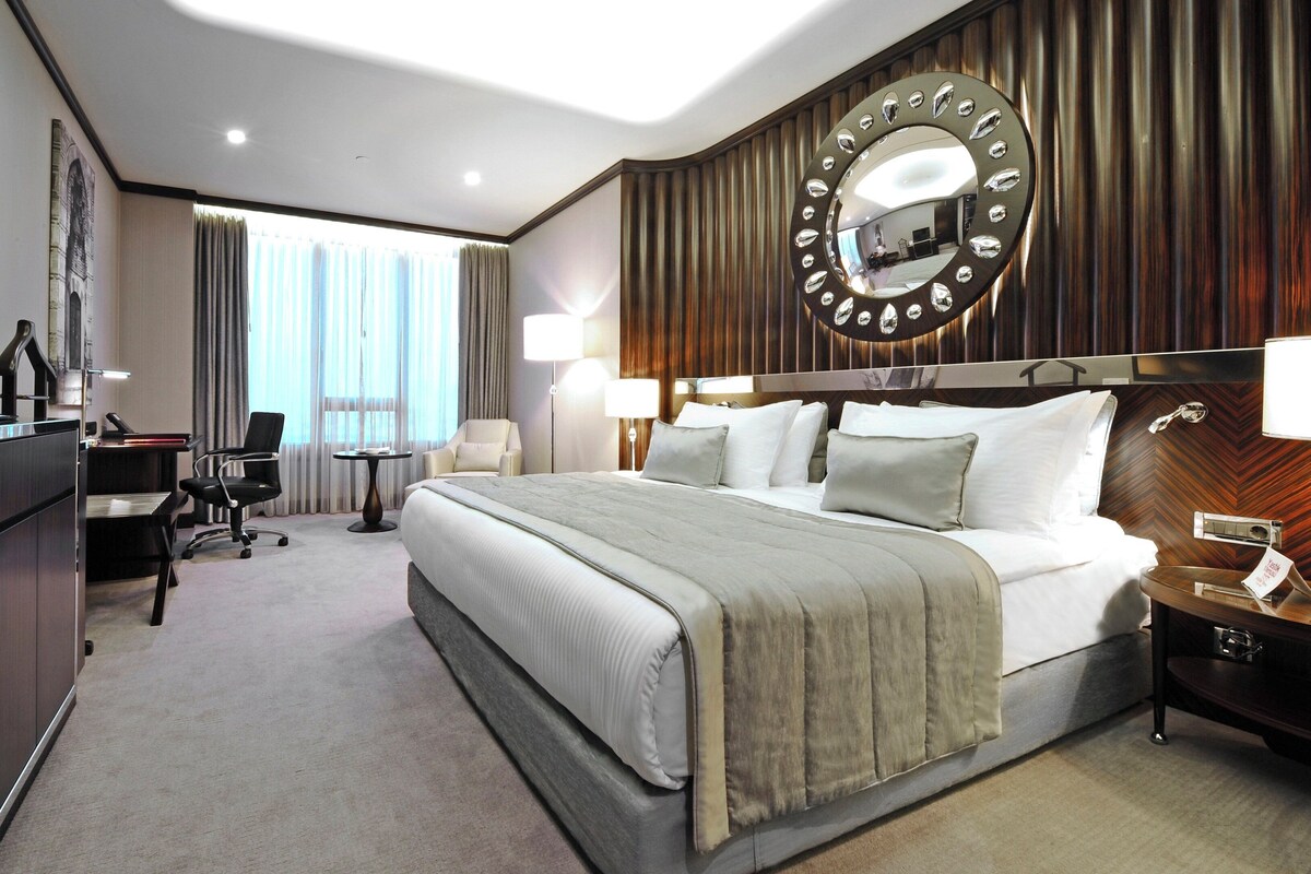 A hotel room with a large bed, a desk with a computer, a TV, and a mirror with a decorative frame.