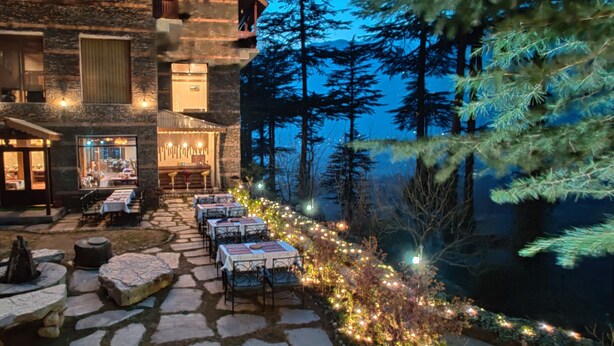 Une terrasse en pierre avec des tables et des chaises, un foyer extérieur et un bâtiment doté de nombreuses fenêtres et d’un espace bar.