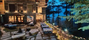 A stone patio with tables and chairs, a fire pit, and a building with multiple windows and a bar area. - Heritage Satikva Resorts (Kullu)