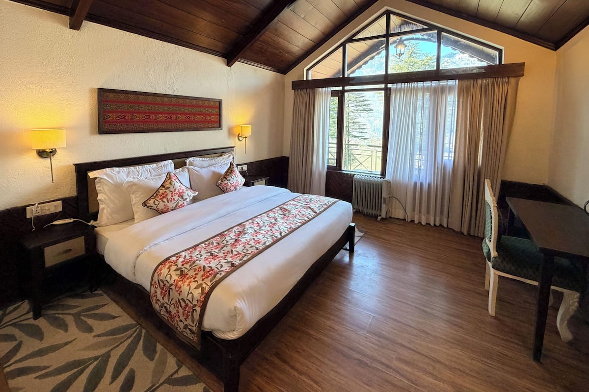 Une chambre d’hôtel avec un grand lit, un bureau, une chaise, une fenêtre avec des rideaux et une lampe fixée au mur.