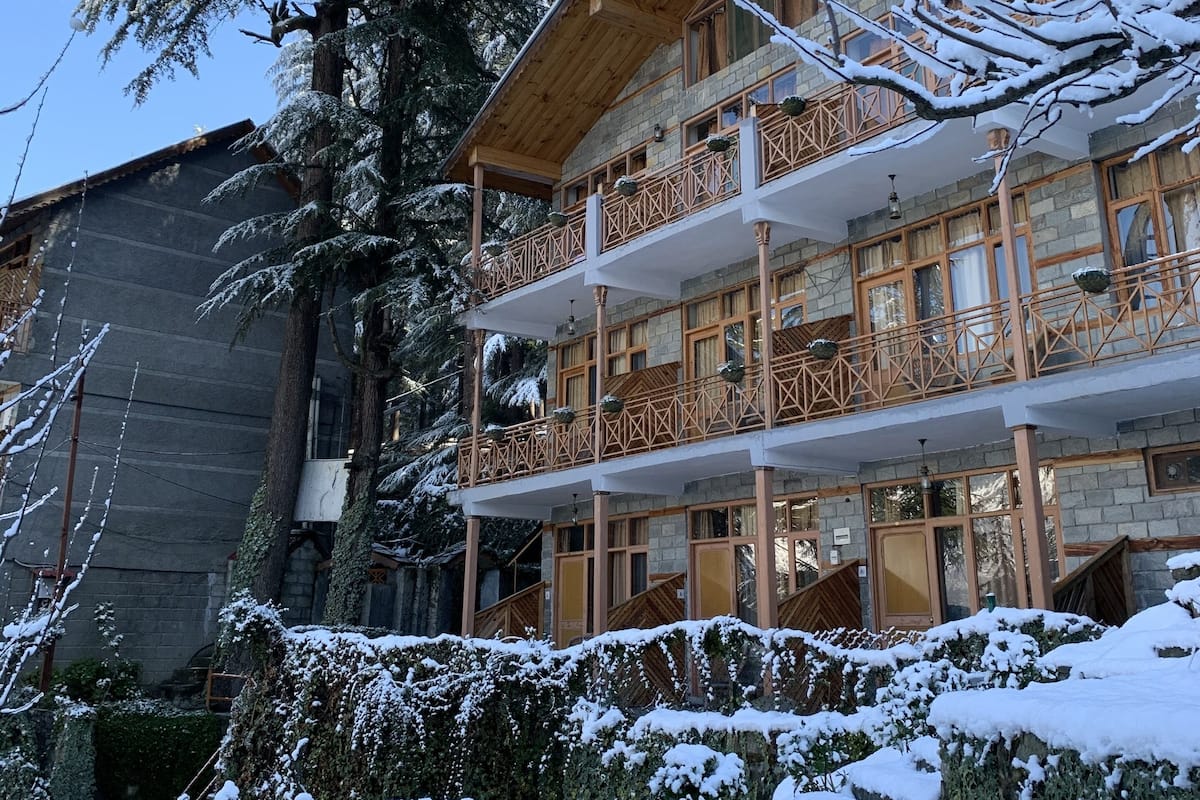 Un immeuble en bois à plusieurs étages, doté de balcons, entouré d’arbres couverts de neige et d’un mur de pierre.