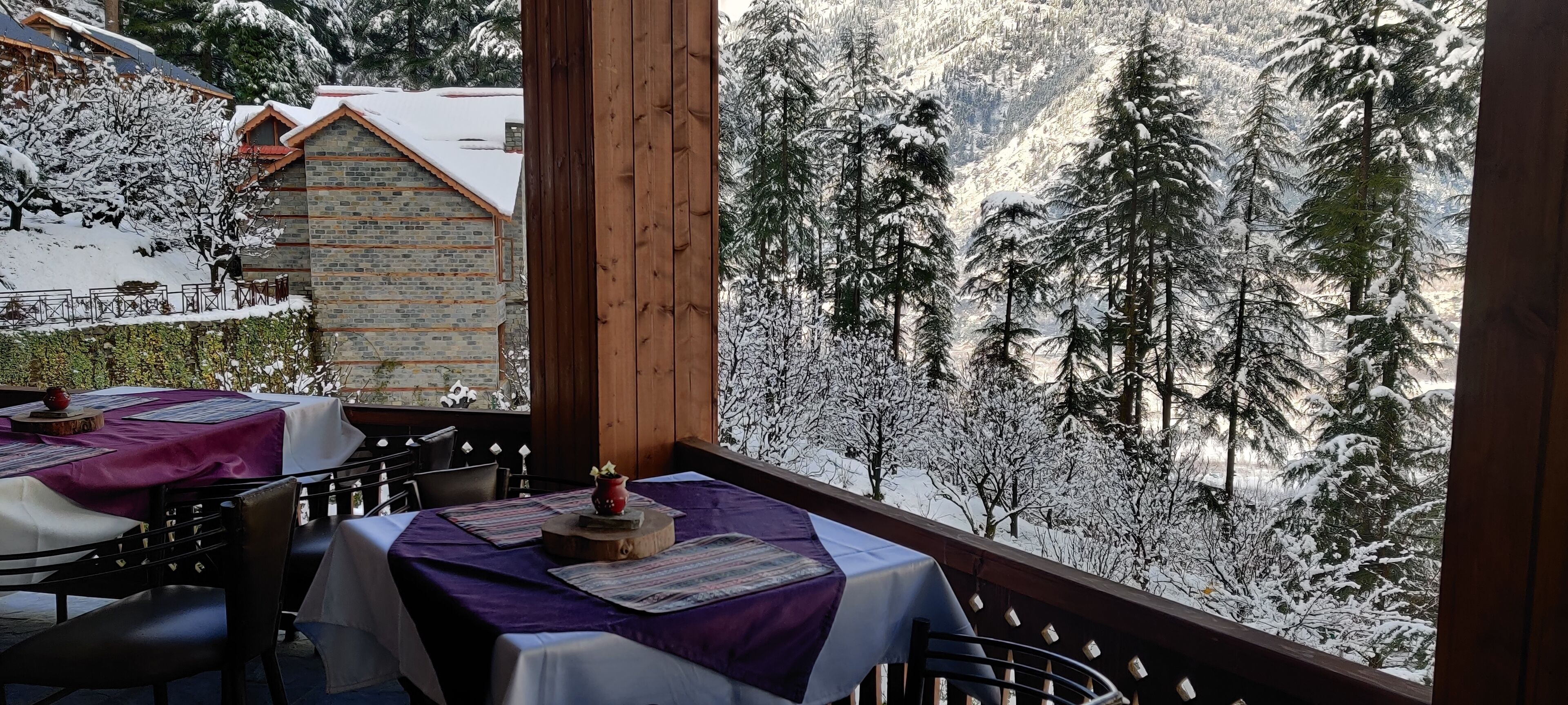 A snow-covered balcony with tables set for dining, overlooking a forested mountain area.