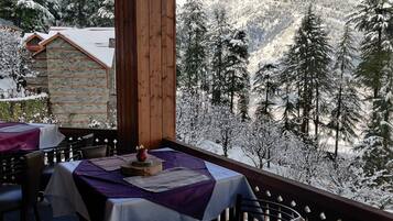 A snow-covered balcony with tables set for dining, overlooking a forested mountain area.