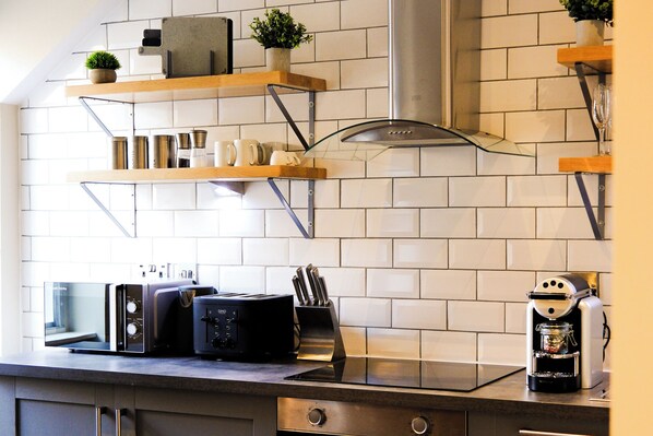 Una cocina con salpicadero de azulejos blancos, estantes de madera, una cafetera, un tostador y un microondas.