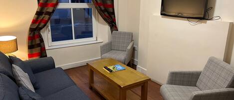 A living room with a sofa, two armchairs, a coffee table, a TV mounted on the wall, and a window with curtains.