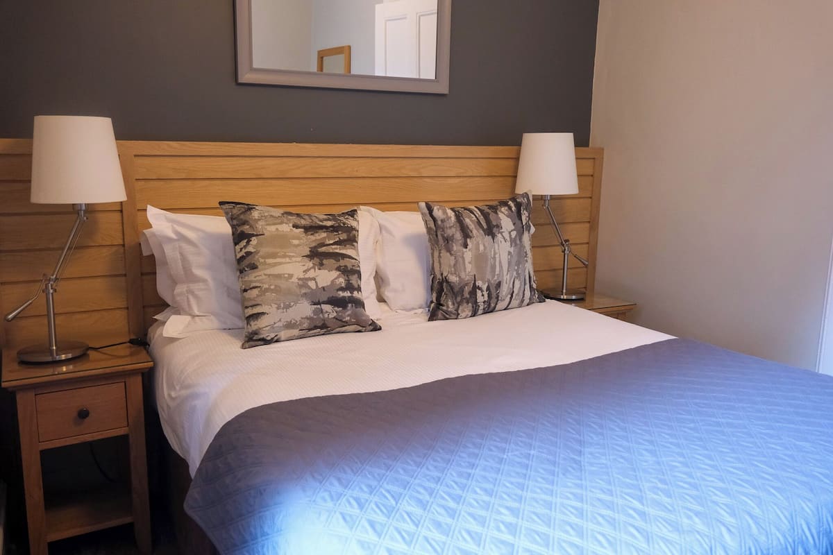 A neatly made bed with a blue quilt, two wooden nightstands with lamps, and a mirror on the wall.