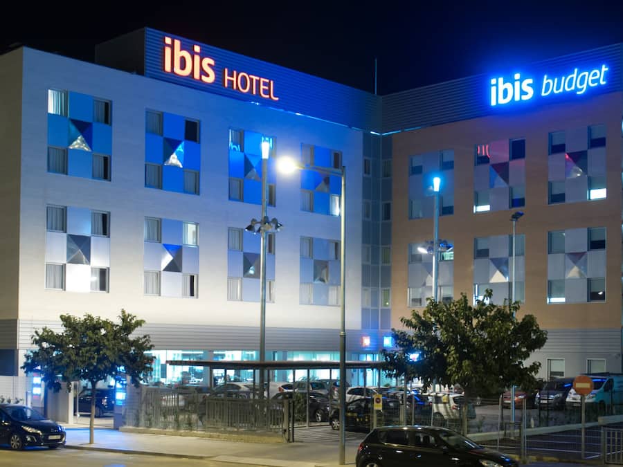 Un hôtel moderne, reconnaissable à son panneau « ibis HOTEL » bien visible, doté d’une section distincte « ibis budget » et d’un parking où sont stationnées plusieurs voitures.