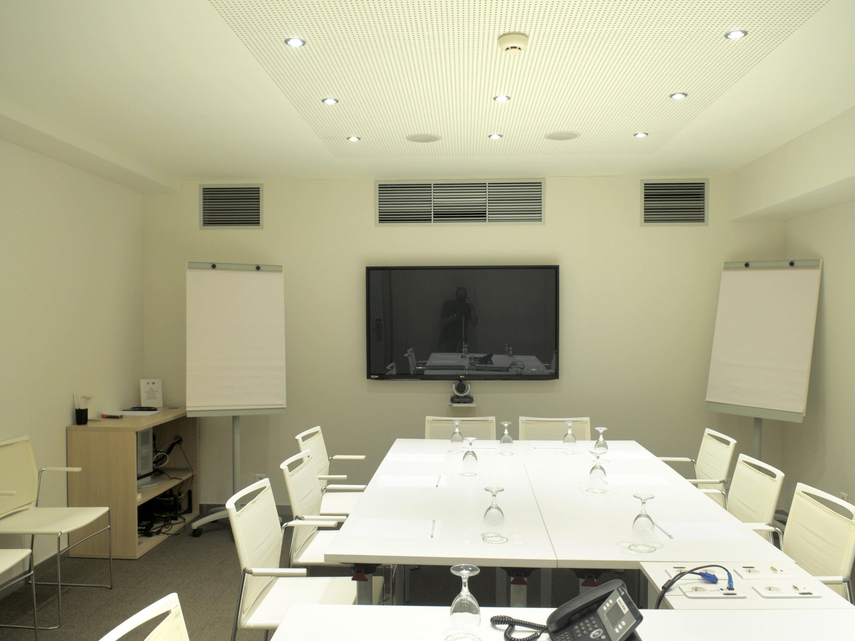 Un salón de conferencias con una mesa larga, sillas blancas, un televisor de pantalla plana, pizarras blancas y un soporte para pizarra.