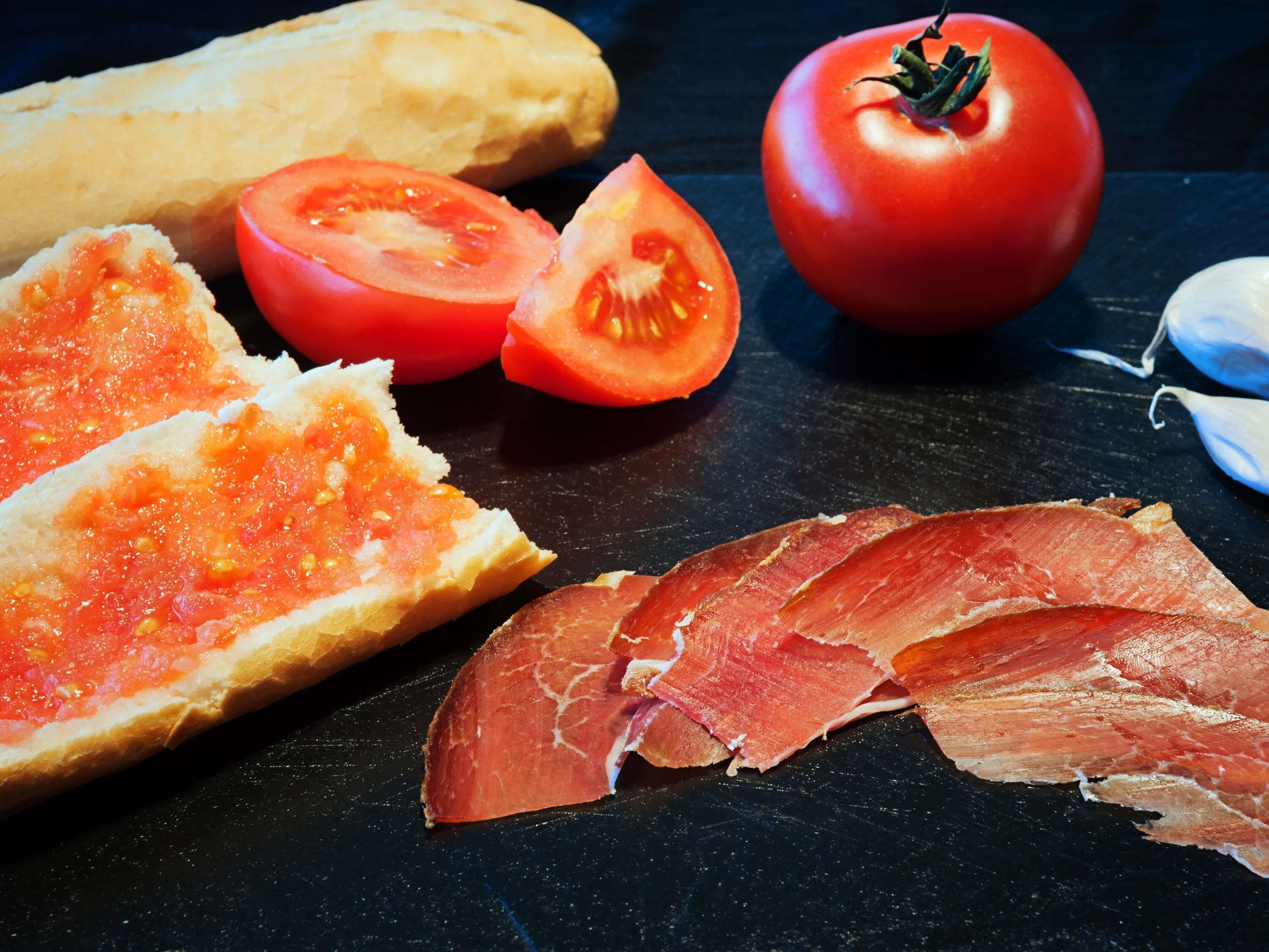 A table with bread, tomato slices, a whole tomato, and sliced meat.