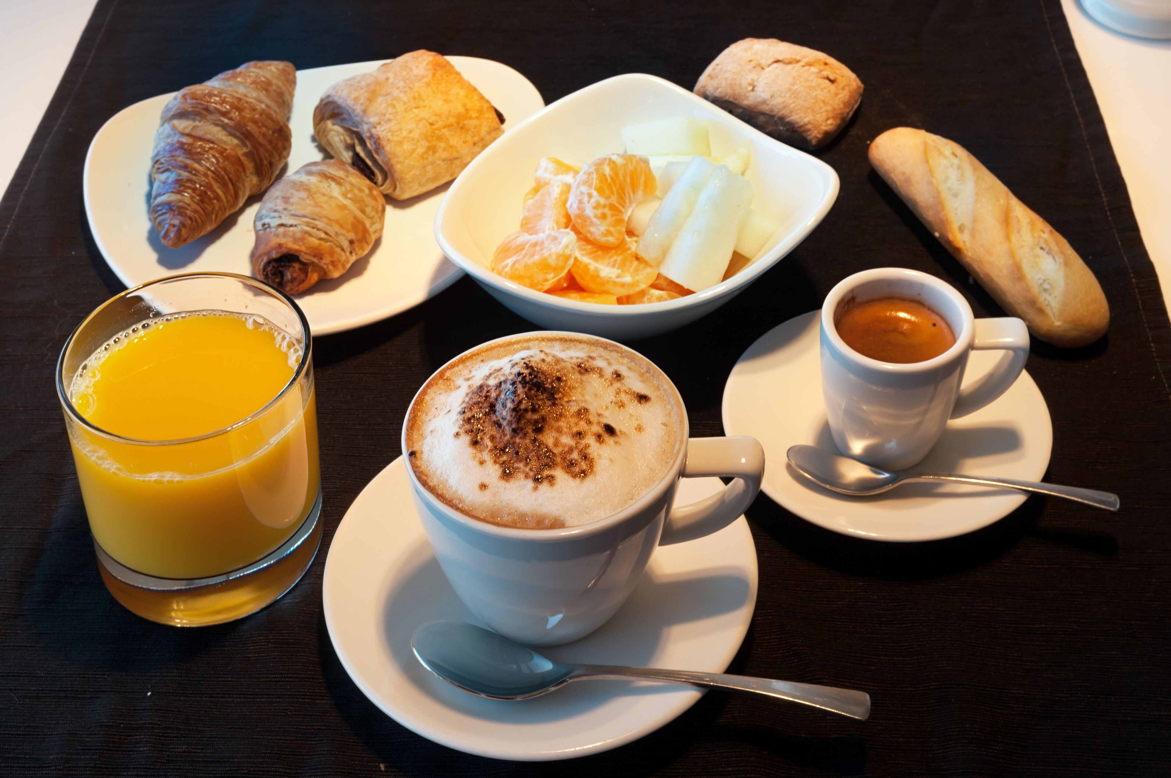 Une table dressée pour le petit-déjeuner avec des croissants, des fruits, un verre de jus d’orange, une tasse de café et une petite tasse d’expresso.