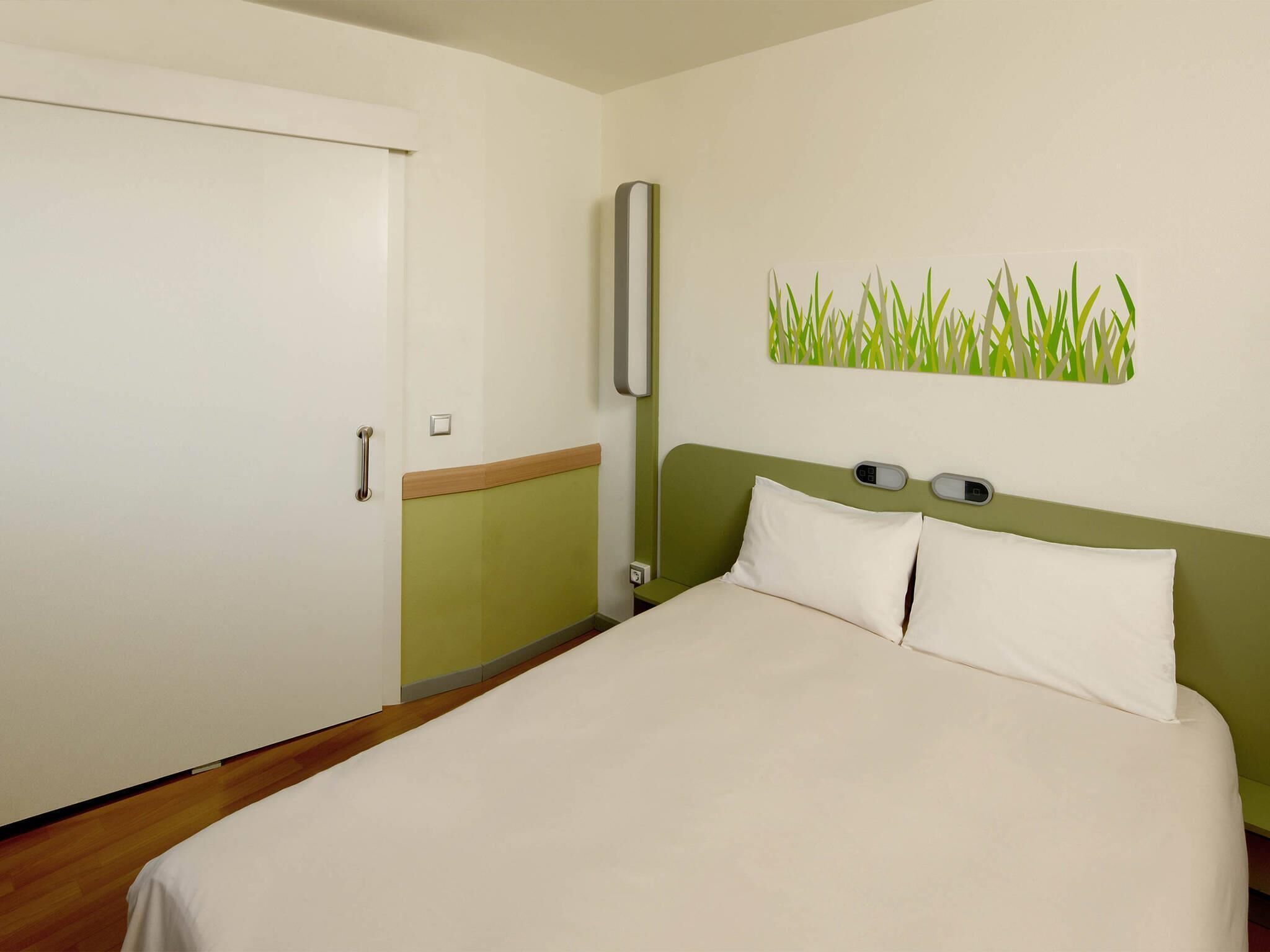 Een hotelkamer met een bed, nachtkastje, een lampje aan de muur en een deur.