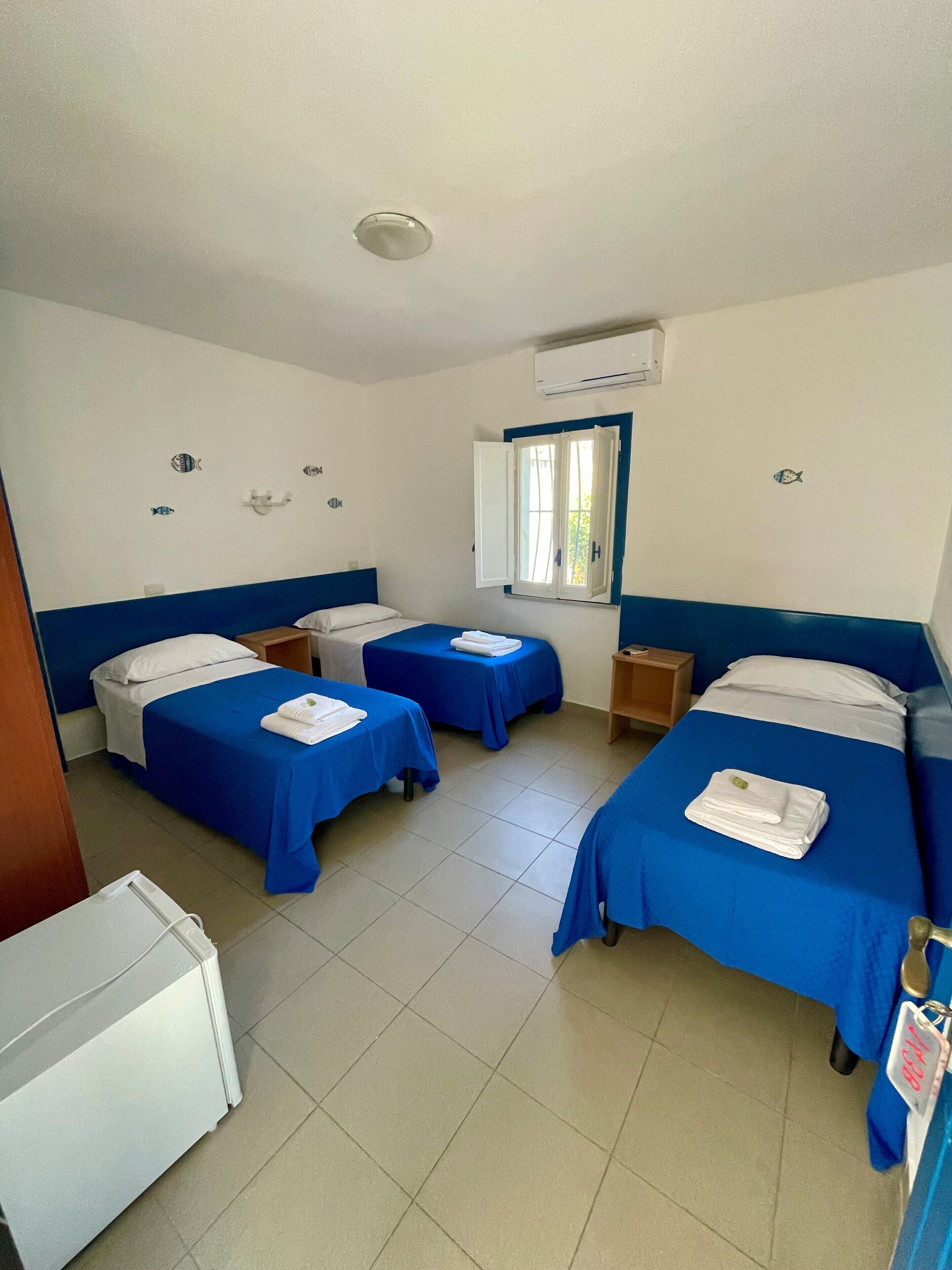 A hotel room with four single beds, a small table, a window with a view, and a white refrigerator.