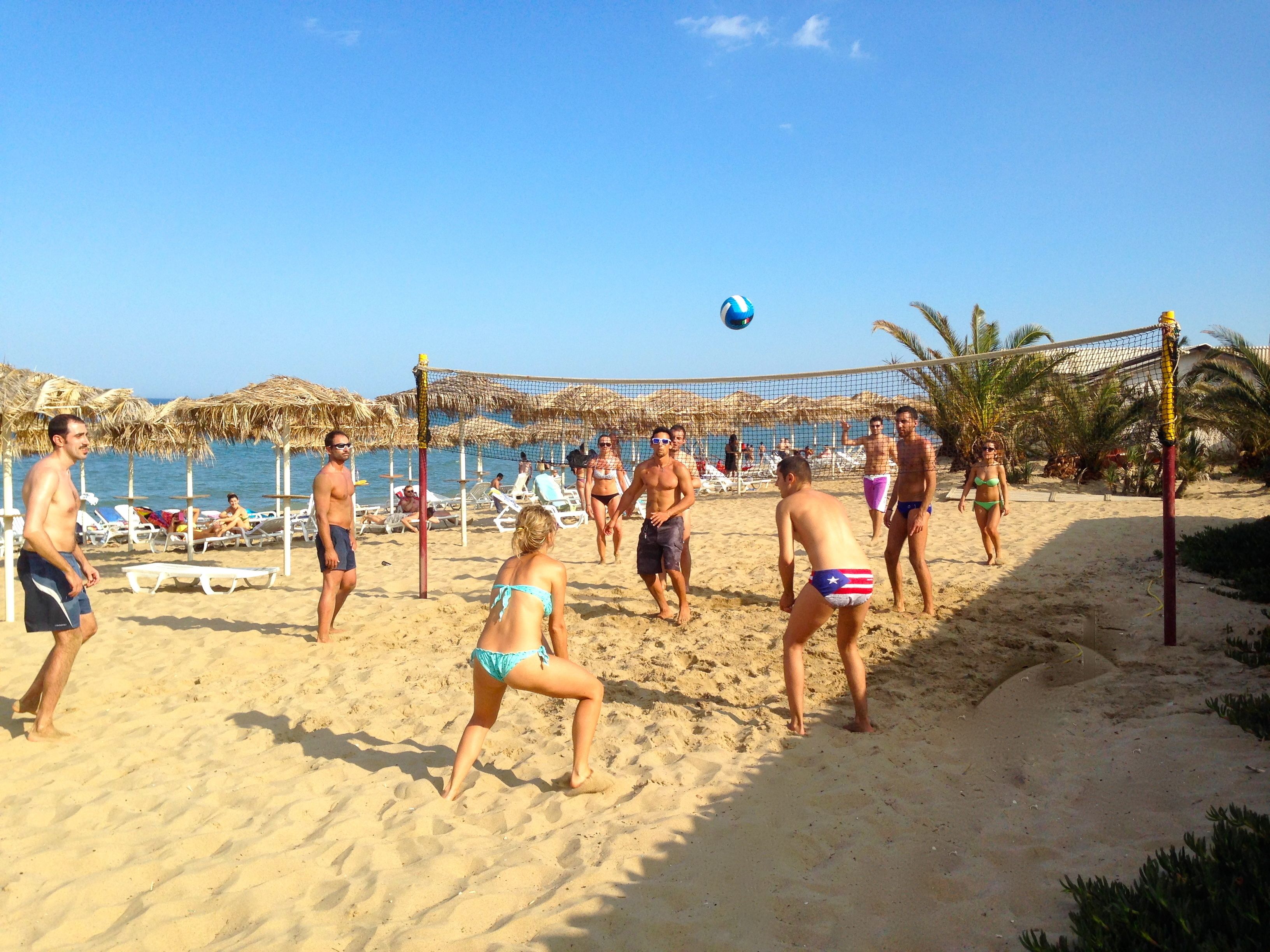 Partido de voleibol de playa con jugadores en traje de baño, una cancha de arena y sombrillas de playa al fondo.