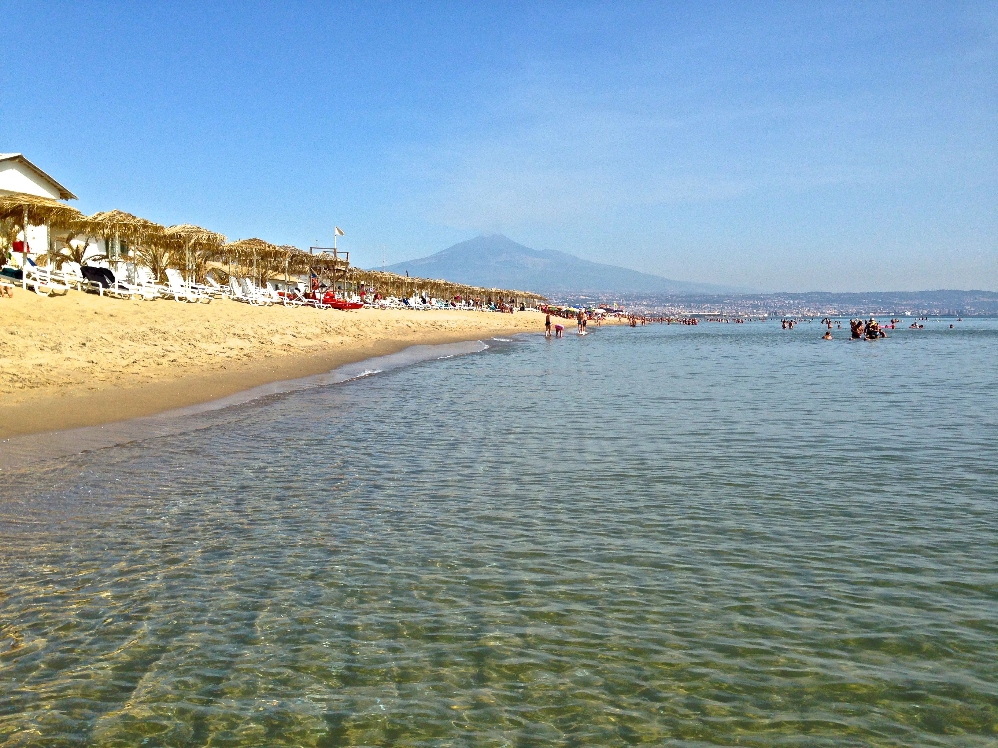 Ein Strand mit klarem, seichten Wasser, einem Sandufer und einem Berg im Hintergrund.