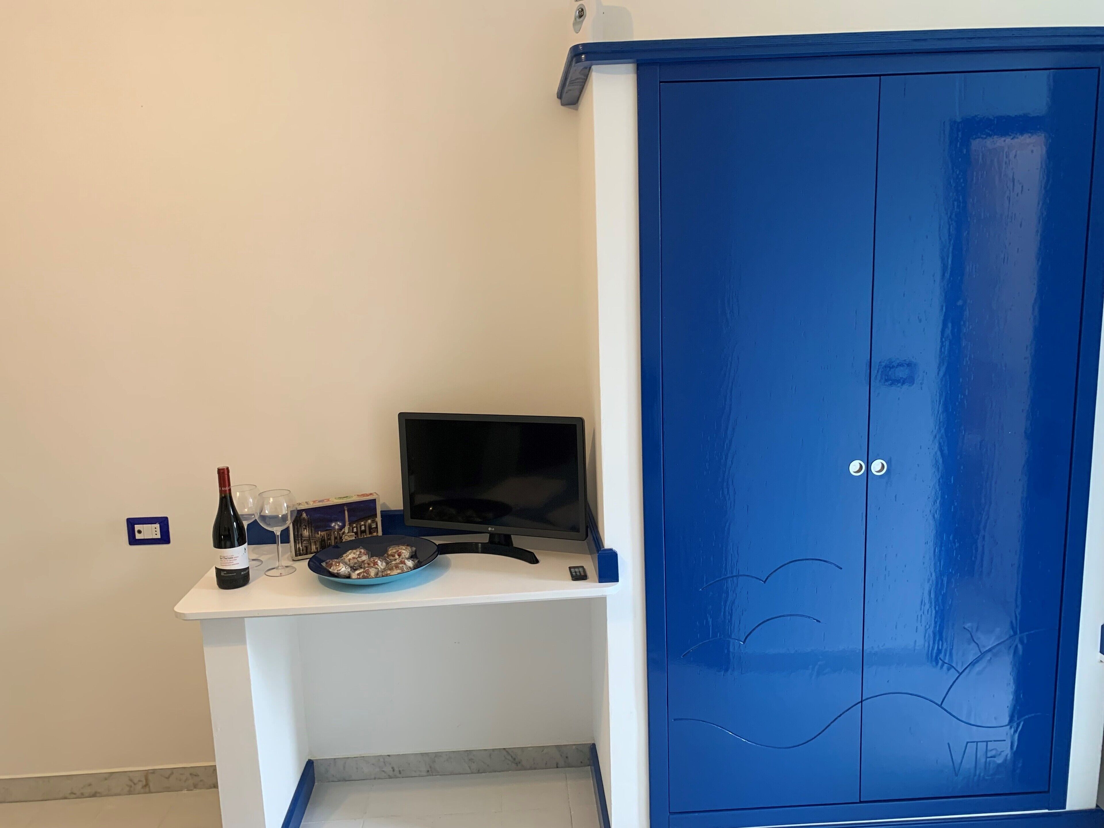 Ein Zimmer mit einem weißen Schreibtisch, einem blauen Kleiderschrank, einem Fernseher, einer Weinflasche und einem Teller Essen.