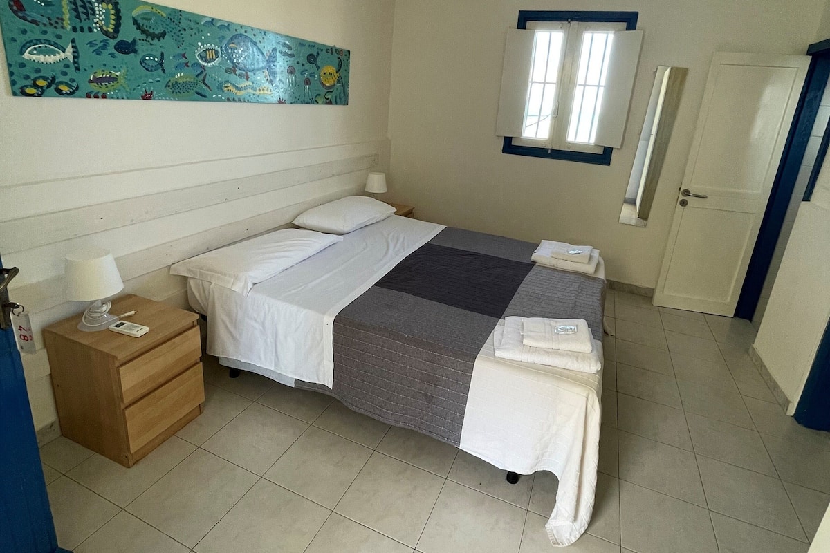 A bedroom with a bed, a nightstand, a wall painting, a window, and a door.