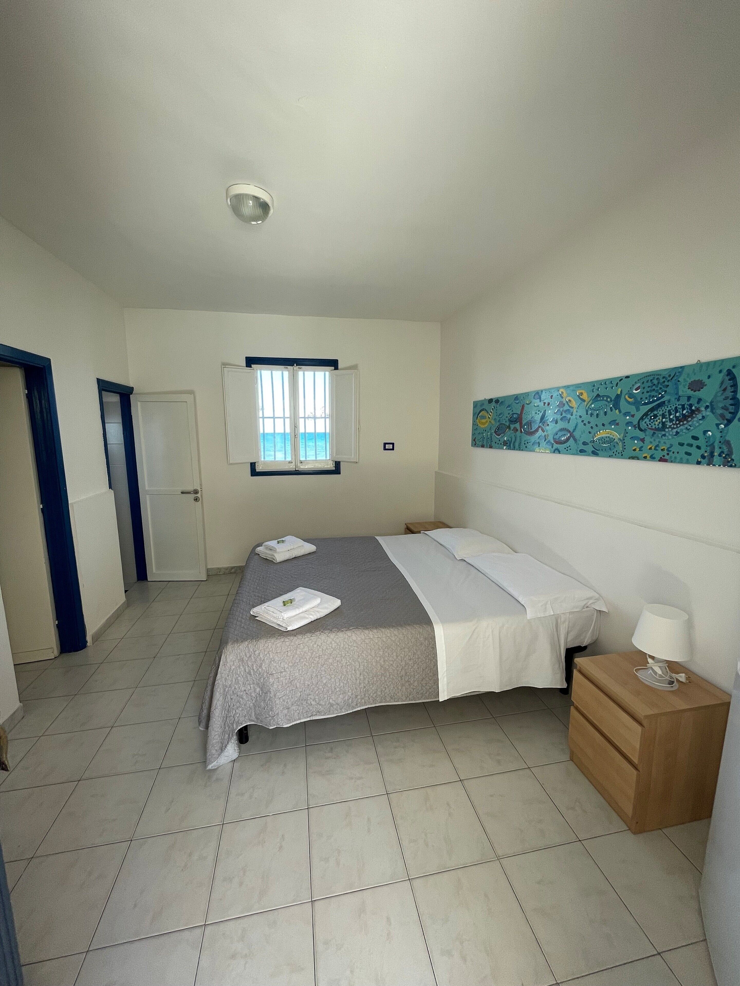 Une chambre à coucher comprenant un lit, une table de chevet, une fenêtre avec vue et une œuvre d’art murale.