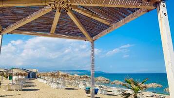 A beach with a wooden pavilion, beach chairs, umbrellas, and a palm tree.
