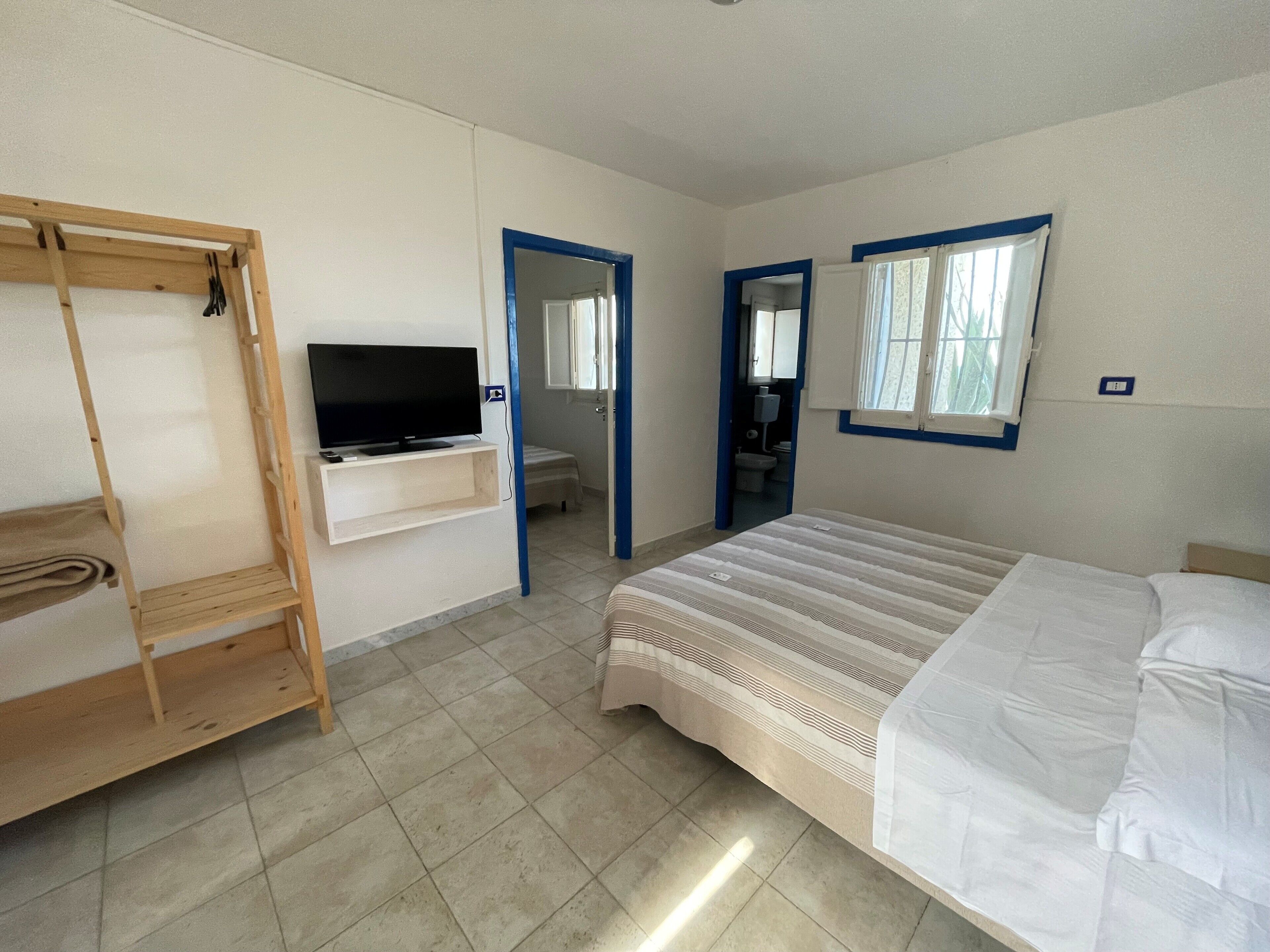 Une chambre à coucher avec un lit, un téléviseur fixé au mur, une étagère en bois et une salle de bain visible à travers une porte ouverte.