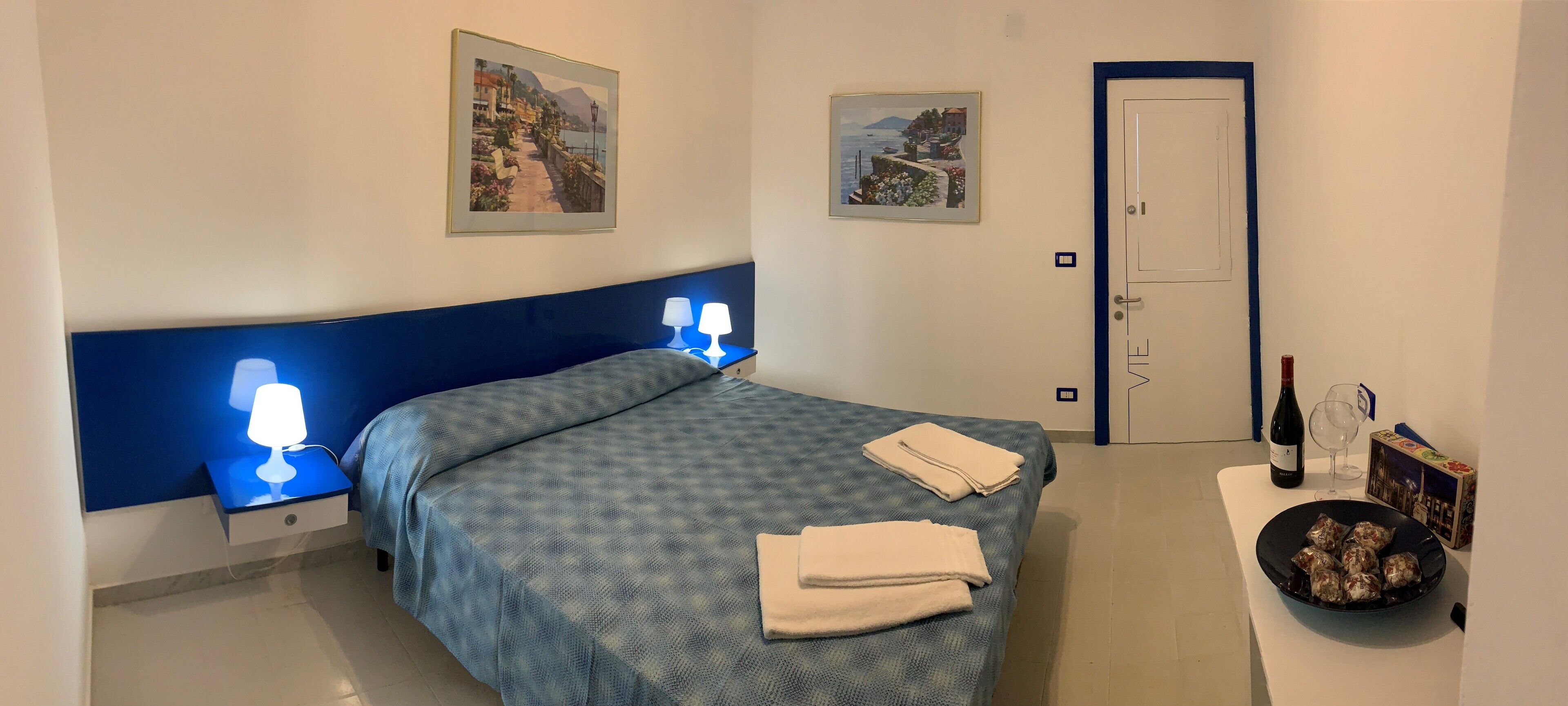 Une chambre à coucher avec un lit, deux lampes fixées au mur, une porte, une petite table sur laquelle se trouvent une bouteille de vin, un verre et une boîte.
