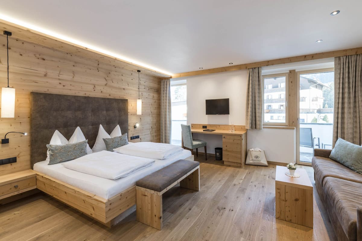 A modern hotel room with a wooden headboard, a bed with pillows, a desk with a chair, a sofa, and a TV mounted on the wall.