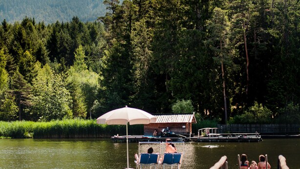 A wooden dock extending into a lake with a row of chairs and an umbrella, surrounded by dense forest and mountains in the background.