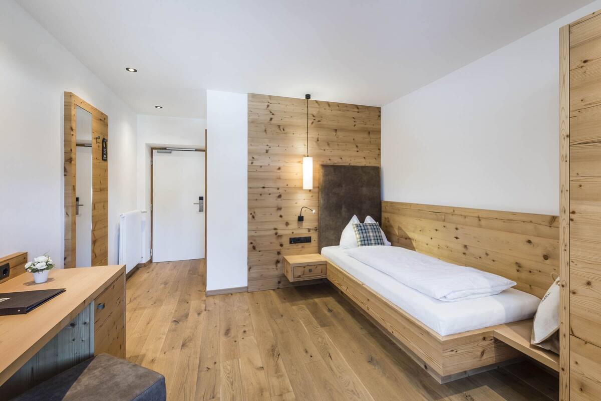 A compact bedroom with a wooden headboard, a bedside table with a lamp, and a small desk area with a plant.