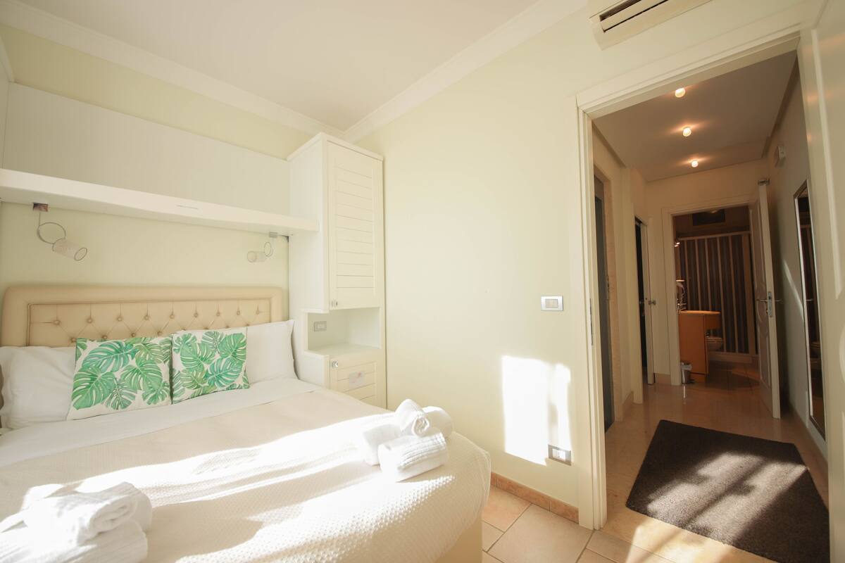 Une chambre d’hôtel comprenant un lit, une table de chevet, une tête de lit, une fenêtre et une porte donnant sur une salle de bain.