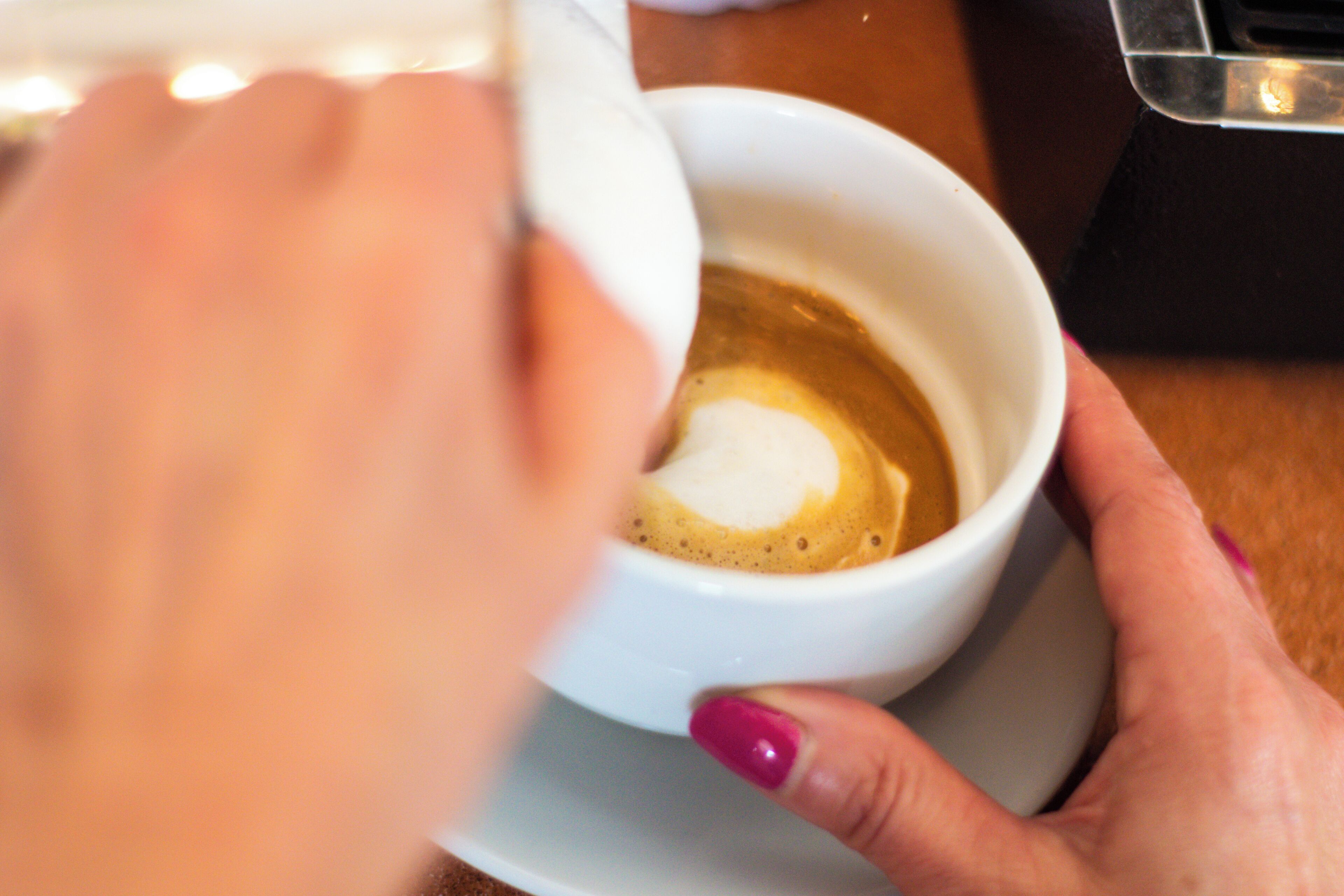 A close-up of a hand holding a white cup with a latte, a saucer, and a spoon.