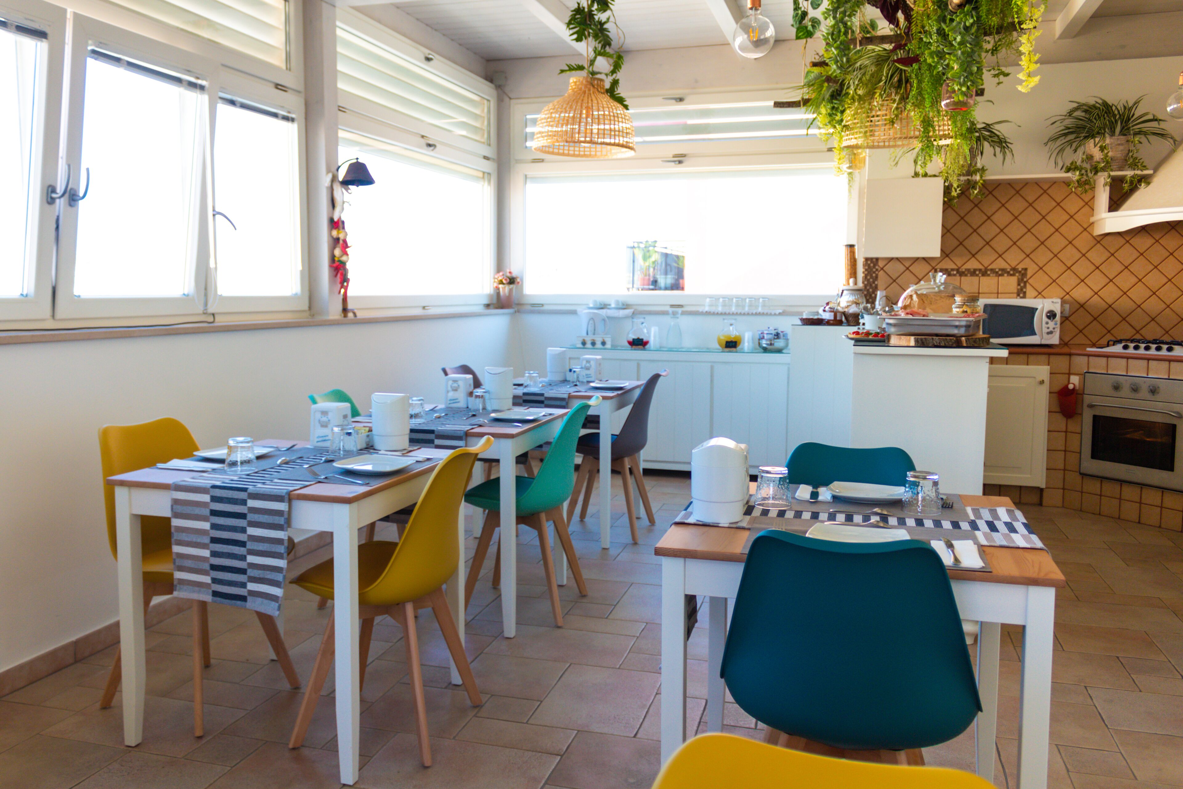A dining area with a table set for four, surrounded by colorful chairs, a kitchen area with appliances, and large windows allowing natural light.