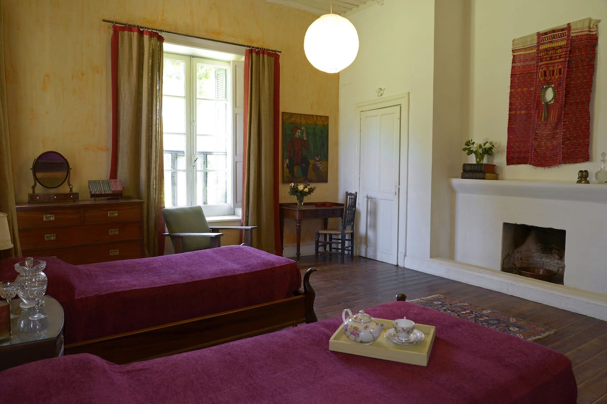 A room with two beds, a fireplace, a dining table with chairs, a desk, a mirror, and a window with curtains.