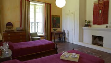 Una habitación con dos camas, chimenea, una mesa de comedor con sillas, escritorio, espejo y ventana con cortinas.