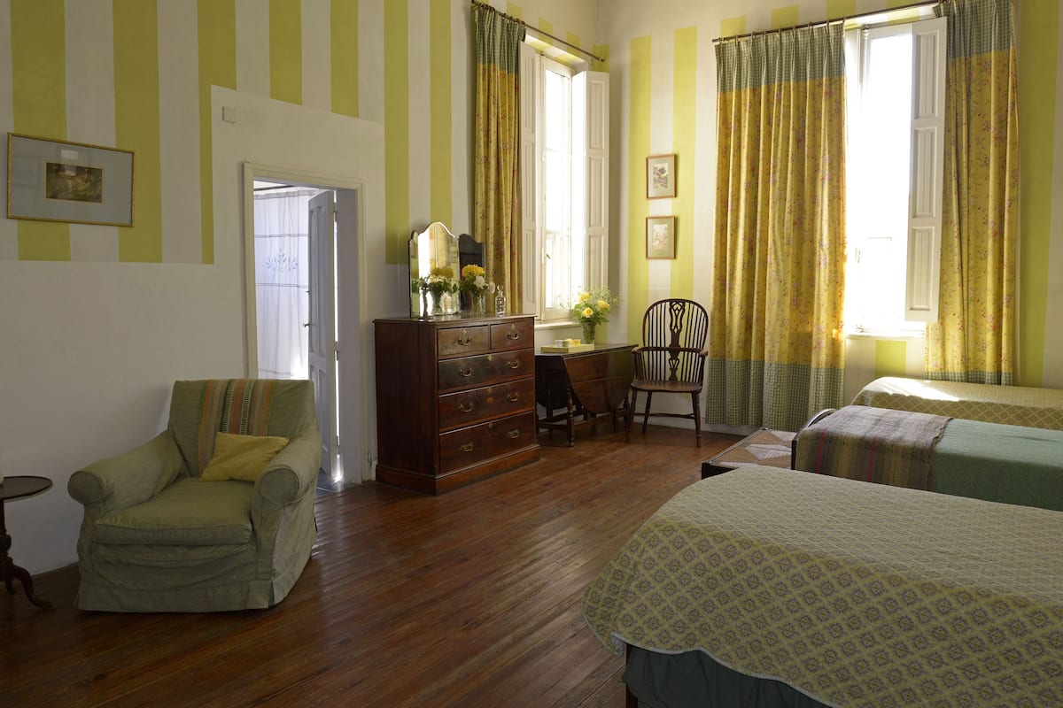 A room with a wooden floor, a green armchair, a wooden dresser, a desk with a chair, and a bed with a patterned bedspread.