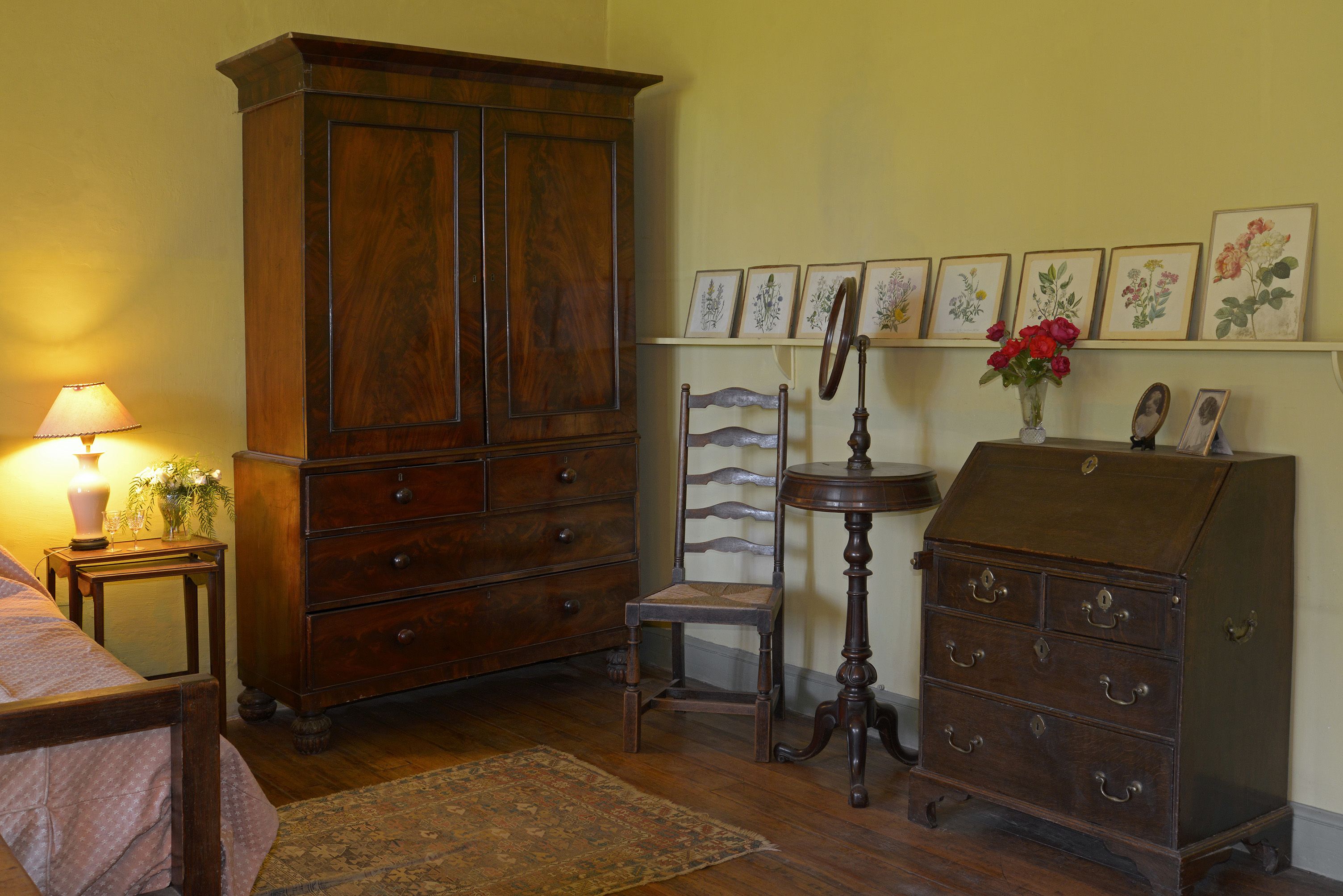 A room with a large wooden cabinet, a chair, a small table with a lamp, and a wall with framed botanical prints.