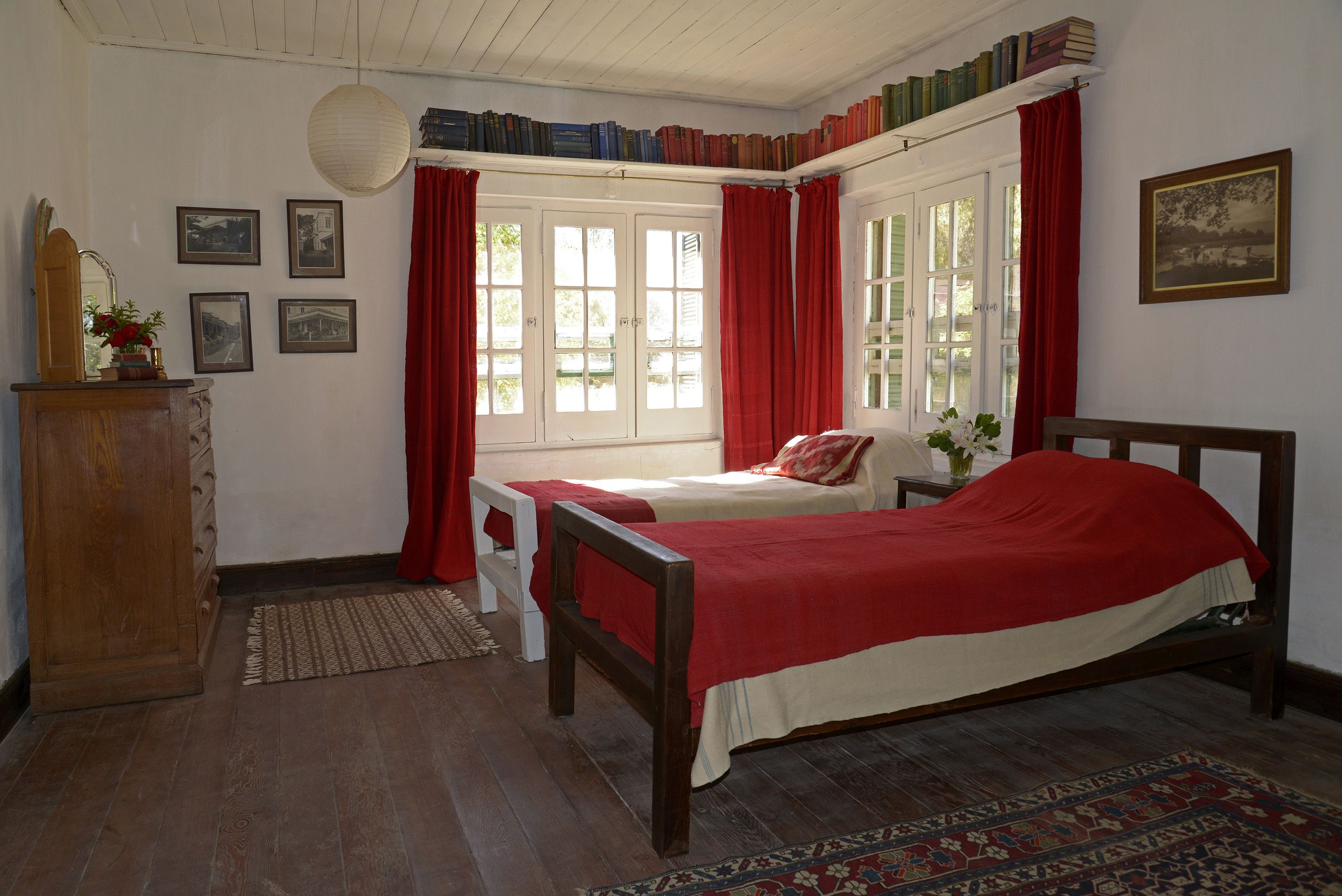 A room with a wooden bed, red curtains, a wooden dresser, and a window with red curtains. There are framed pictures on the wall and a bookshelf with books.