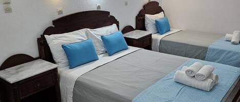 Une chambre d’hôtel avec deux lits, chacun doté d’une tête de lit en bois, de draps blancs et d’oreillers bleus. On y trouve des appliques murales et une petite table de chevet avec un tiroir.