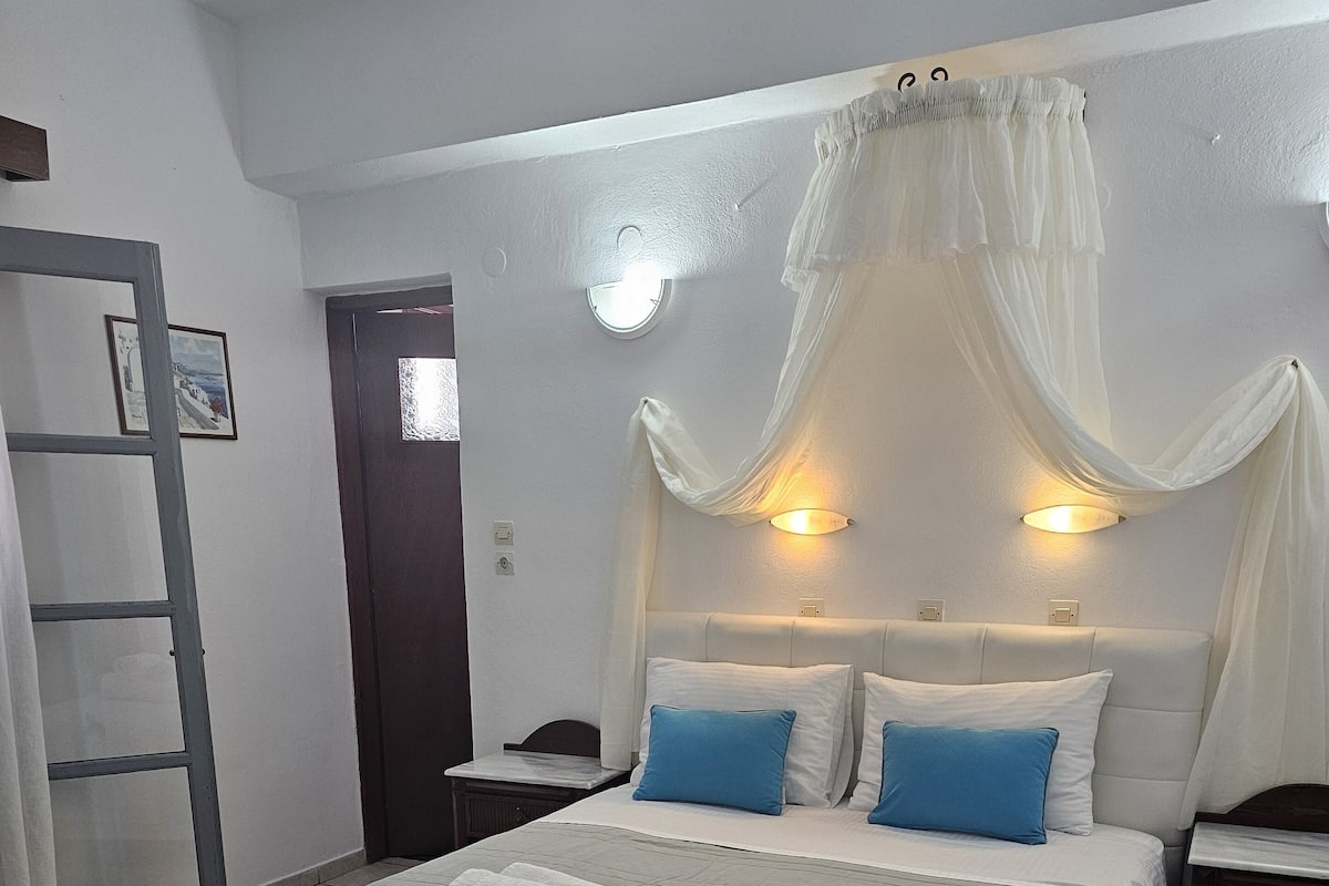 Ein ordentlich bezogenes Bett mit einer blauen, gemusterten Tagesdecke und zwei Kissen, ein Nachttisch mit einer Lampe sowie eine Tür mit Vorhang.