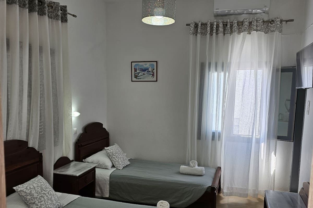 Ein Hotelzimmer mit zwei Einzelbetten, einem Holztisch, einem Stuhl, einem kleinen Tisch, einem an der Wand befestigten Fernseher und einer Klimaanlage.