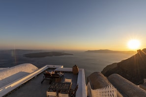 A terrace with a table and chairs overlooking a body of water and distant islands during sunset. - Folia Bianca Apartments (Santorini)