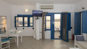 A room with a TV, a dining table, and a view of the sea through open doors. - Folia Bianca Apartments (Santorini)