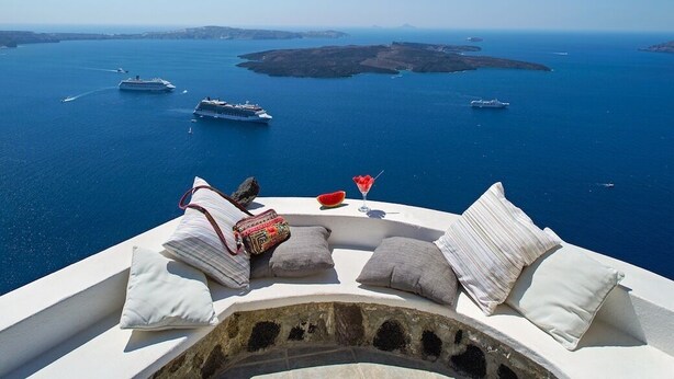 A seaside terrace with a view of the sea and boats, featuring a white cushioned seating area with pillows and a glass of drink. - Folia Bianca Apartments (Santorini)