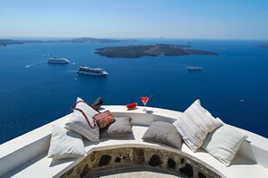 Terraza frente al mar con vista al océano y barcos, que incluye una zona de estar blanca con cojines y una bebida en un vaso.