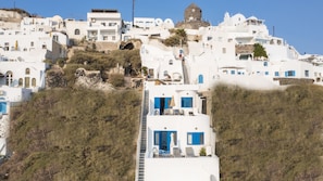 A white building with blue doors and windows, surrounded by greenery and other white buildings. - Folia Bianca Apartments (Santorini)
