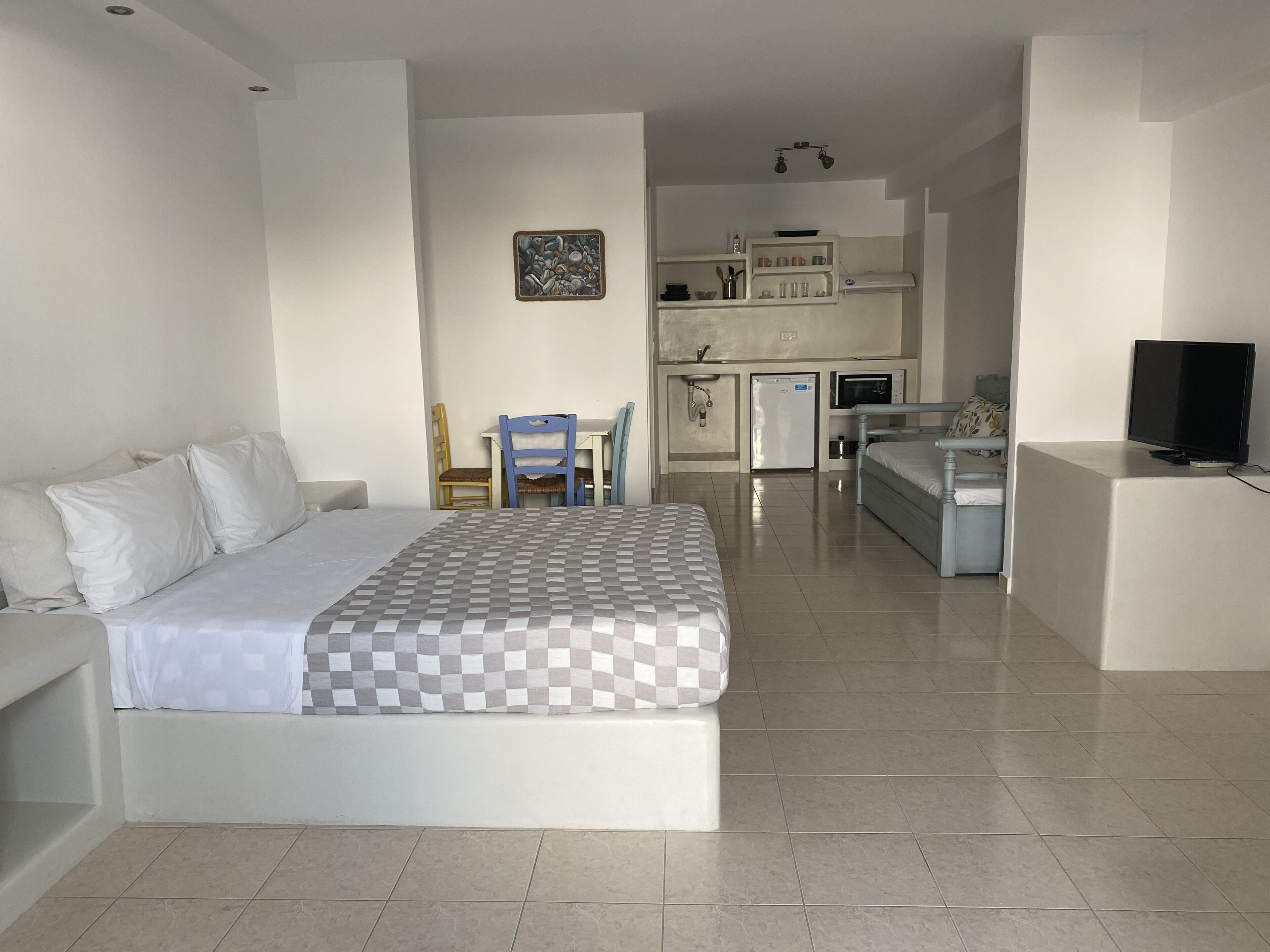 A bedroom with a bed, a desk, a chair, a television, and a kitchenette.